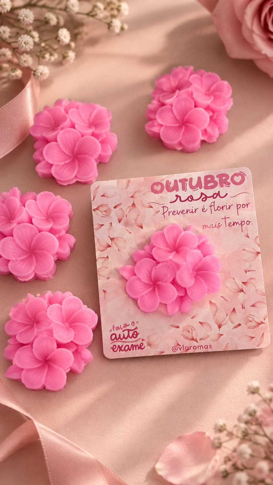 Sabonetes artesanais em formato de flor rosa da campanha Outubro Rosa da VL Aromas, ao lado de cartão “Prevenir é florir por mais tempo”, sobre tecido rosê com fita de cetim