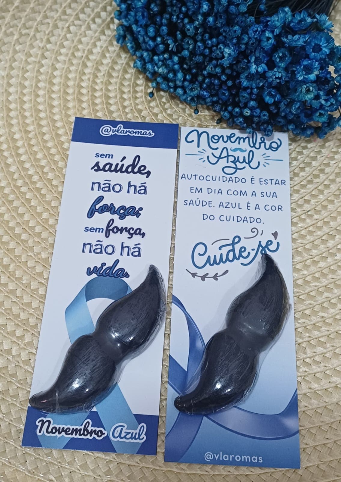 Marcadores de página da campanha Novembro Azul da VL Aromas, com sabonete artesanal em formato de bigode e mensagens de autocuidado masculino, sobre sousplat de palha com flores azuis